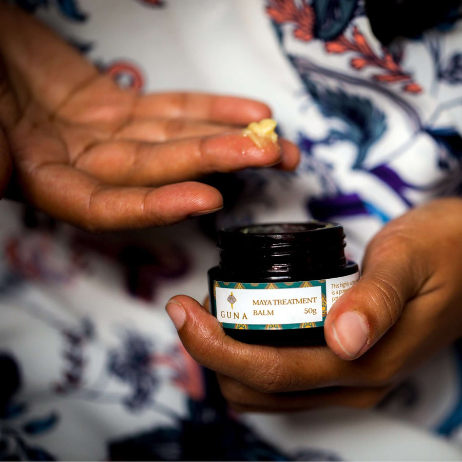 An Indian woman holding an open jar of Guna's Maya Treatment Balm in one hand, scooping out a pea-sized amount in her finger