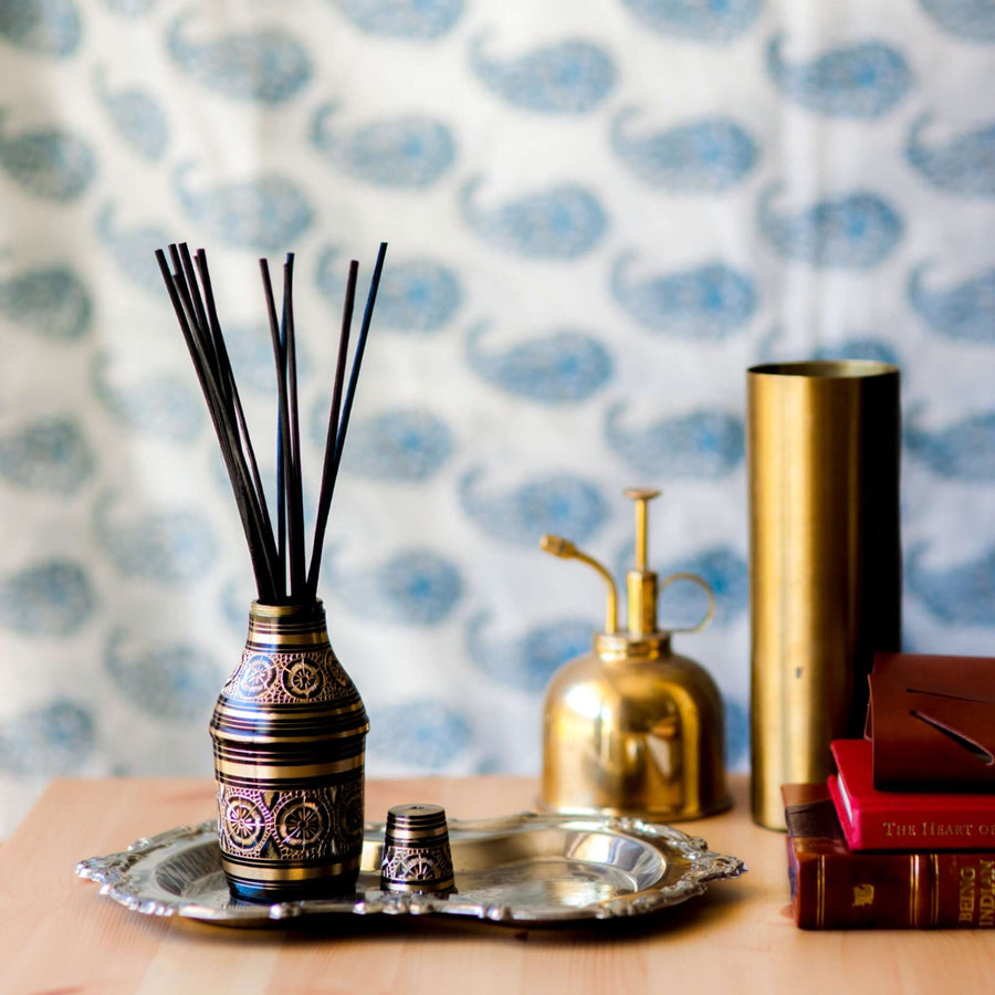 Guna's brass diffuser bottle with black reeds sitting on a silver tray, with leather bound books and a brass vase in the background, and an out of focus white and blue paisley sari in the background