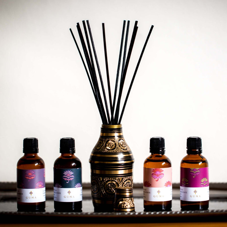 Guna's brass diffuser bottle with black reeds on a silver tray, with 50ml bottles of Guna's Diffuser Oils lined up next to it